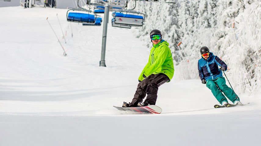 Skier and Rider Make Turns Under Chairlift on Snowy Day at Mount Snow
