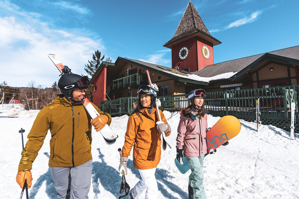 Friends Carrying Equipment Walk on Snow with Clocktower in Background at Mount Snow
