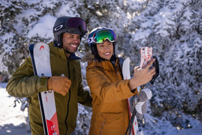 Couple Holding Skis Take Selfie in front of Snowy Trees at Mount Snow Summit