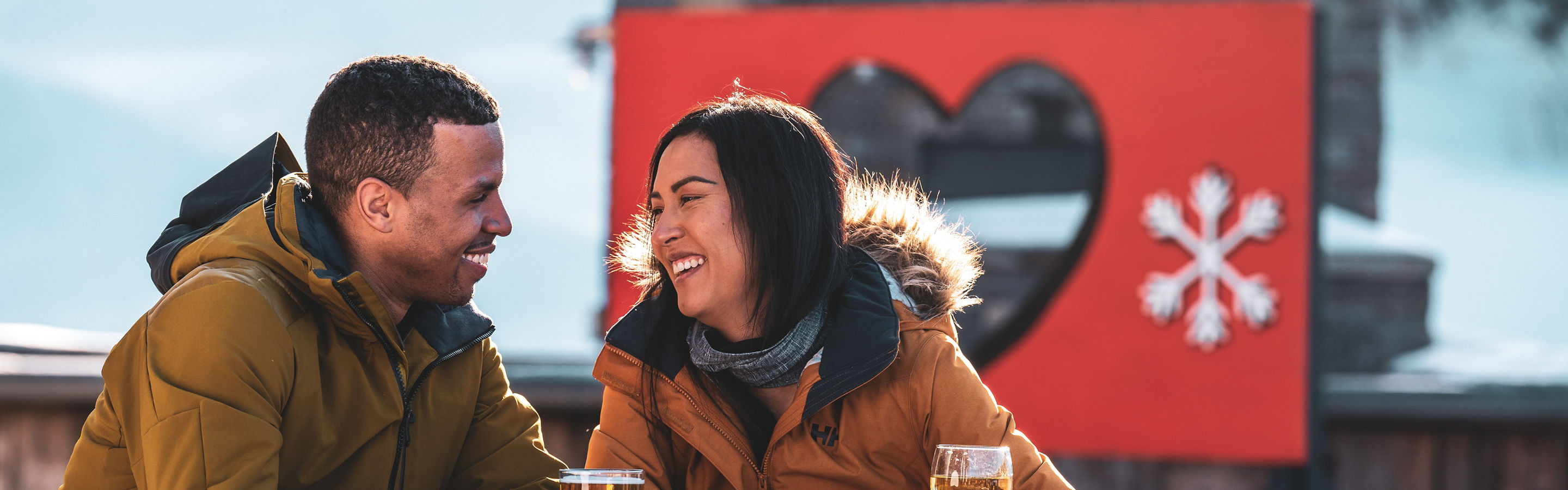 Couple Smiles over Drinks on Carinthia Base Lodge Deck in front of We Love Snow Sign at Mount Snow