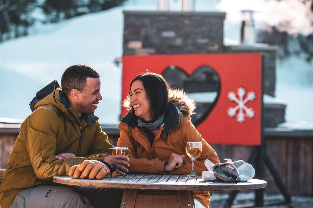 Couple Smiles over Drinks on Carinthia Base Lodge Deck in front of We Love Snow Sign at Mount Snow