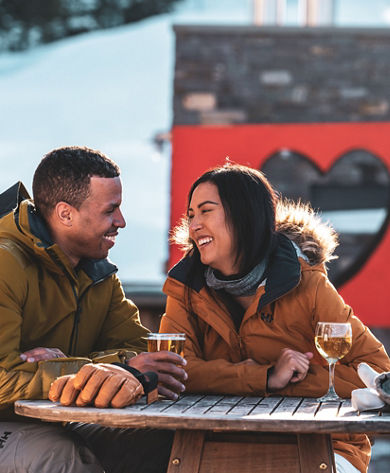 Couple Smiles over Drinks on Carinthia Base Lodge Deck in front of We Love Snow Sign at Mount Snow