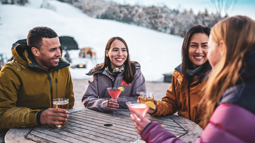 Friends Smile Over Drinks on Carinthia Base Lodge Deck  with Trails in Background at Mount Snow