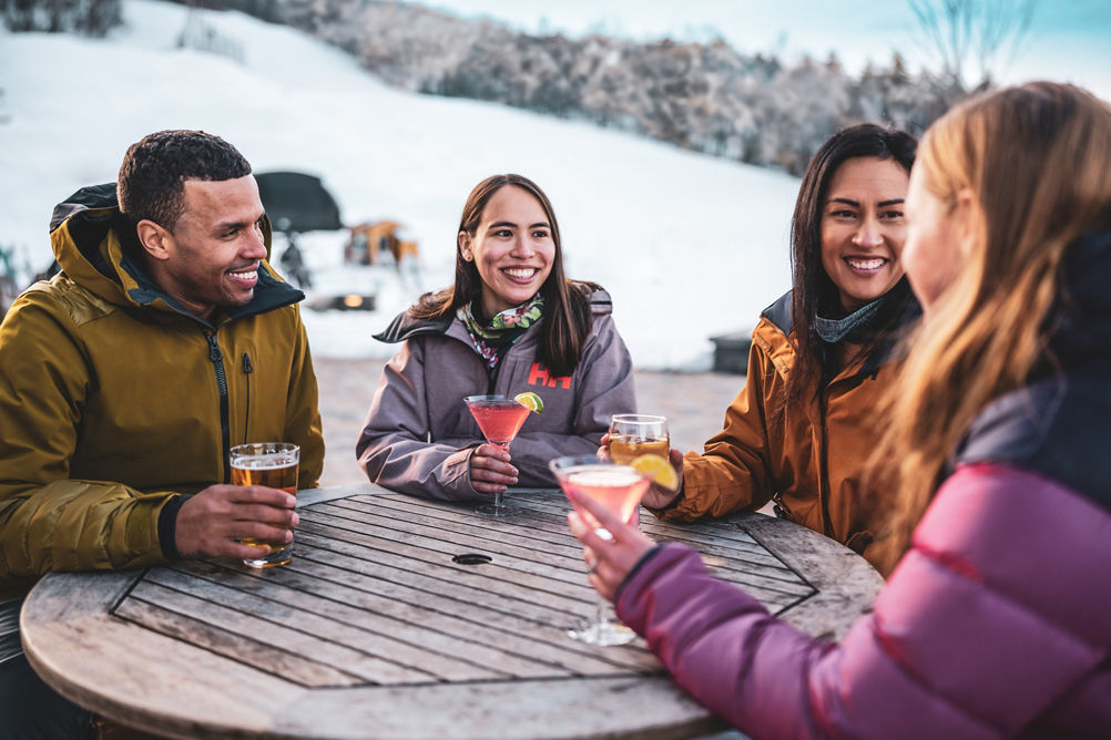 Friends Smile Over Drinks on Carinthia Base Lodge Deck  with Trails in Background at Mount Snow