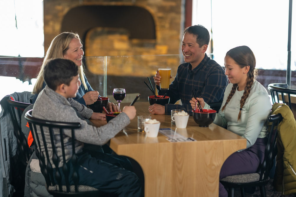 Family Eating Ramen Bowls, Kids with Hot Chocolate, Parents with Wine and Beer in with C on Fireplace in background in Carinthia Base Lodge at Mount Snow
