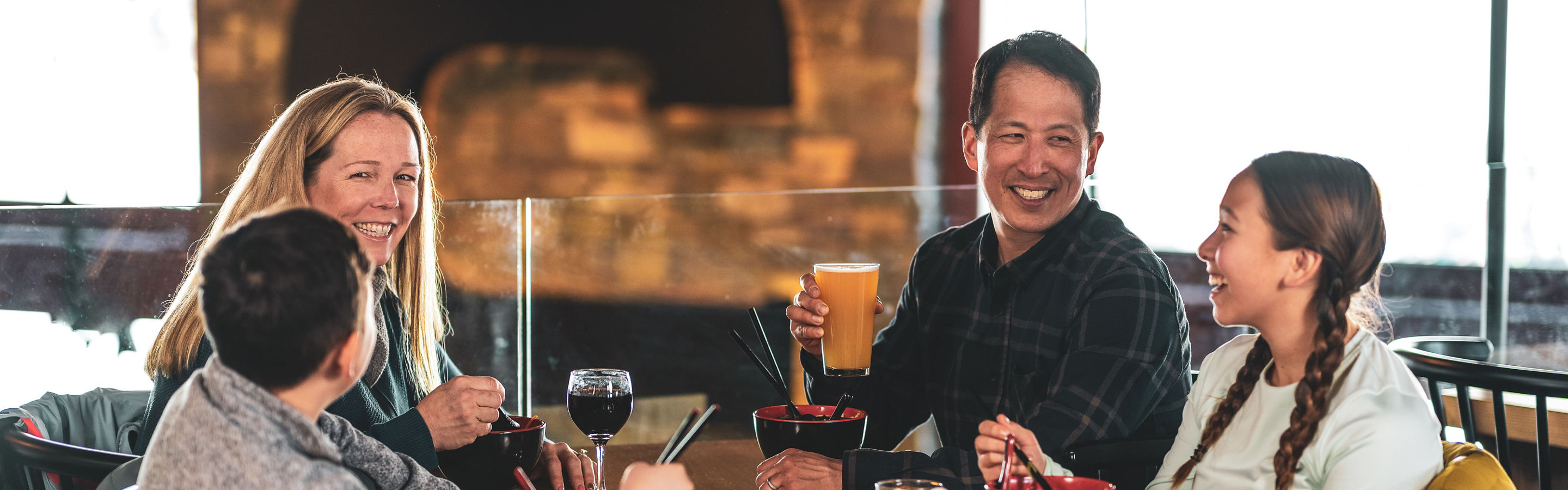 Family Eating Ramen Bowls, Kids with Hot Chocolate, Parents with Wine and Beer in with C on Fireplace in background in Carinthia Base Lodge at Mount Snow