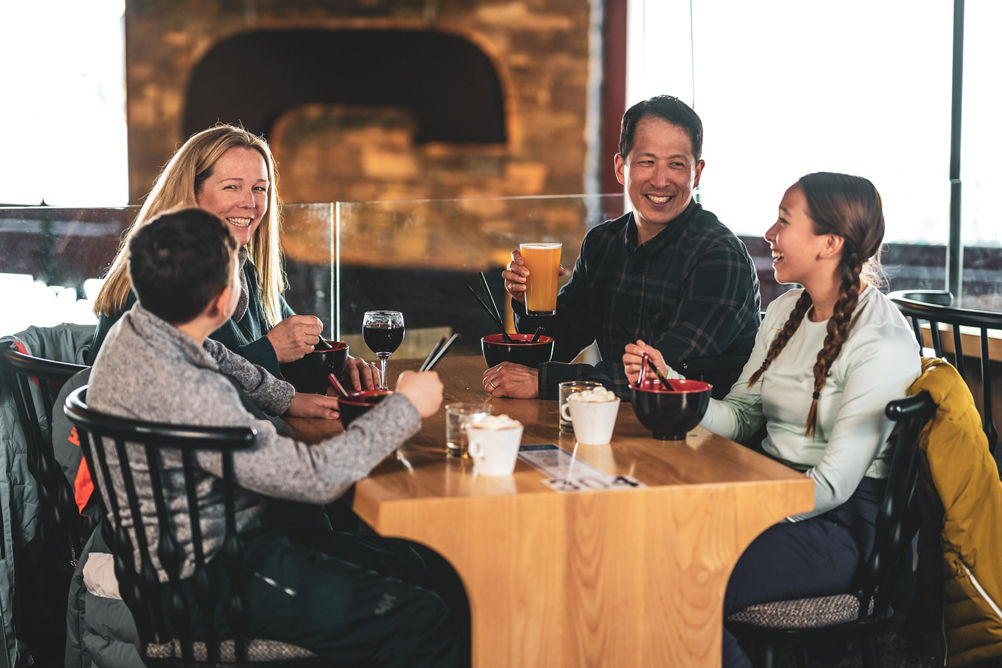 Family Eating Ramen Bowls, Kids with Hot Chocolate, Parents with Wine and Beer in with C on Fireplace in background in Carinthia Base Lodge at Mount Snow