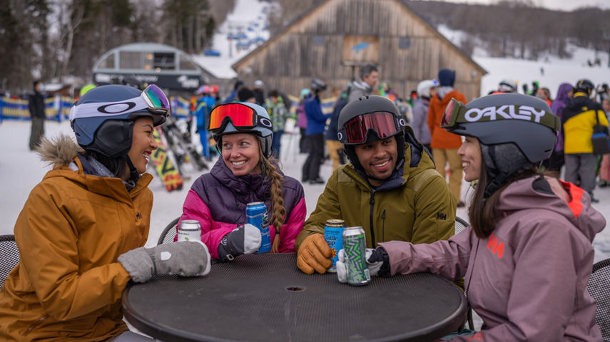Friends Sit Around Table and Smile with Canned Beers in Main Base Area with Bluebird Barn in Background at Mount Snow