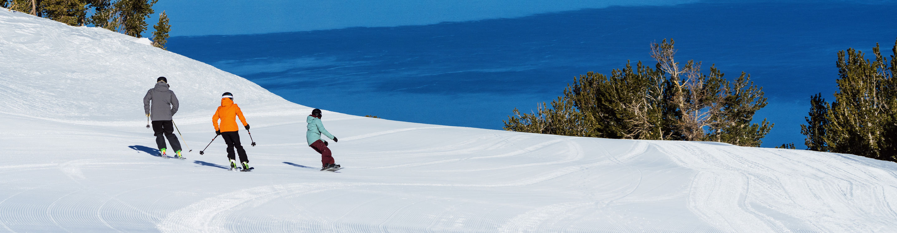 Friends Ski and Snowboard in Unison Down a Run at Heavenly with a Scenic View of Lake Tahoe in the Background